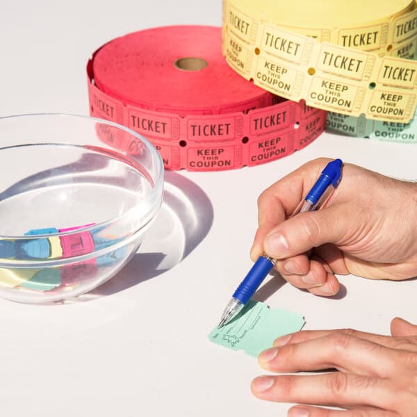 A person using a blue pen to write on a green Carnival King raffle ticket next to a bowl of water.