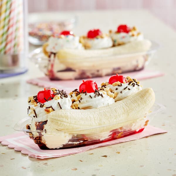 A clear plastic banana split boat filled with a banana split dessert, topped with whipped cream, cherries, nuts, and chocolate sprinkles.