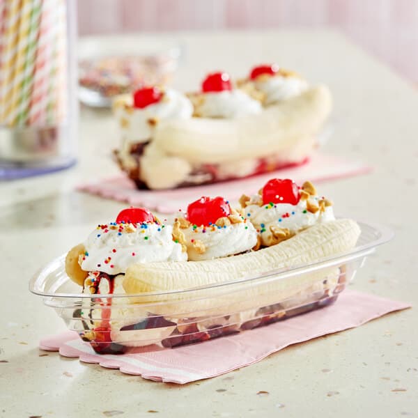 A clear plastic banana split boat filled with a banana split dessert, topped with whipped cream, cherries, and sprinkles.