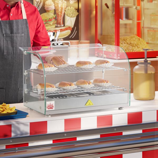 A man wearing a red shirt and apron standing behind a countertop with hot dogs in a glass display case.