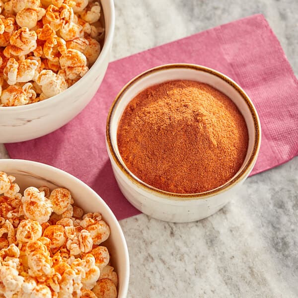 A bowl of popcorn with red and yellow powder next to a bowl of Carnival King Smokehouse Popcorn Seasoning.